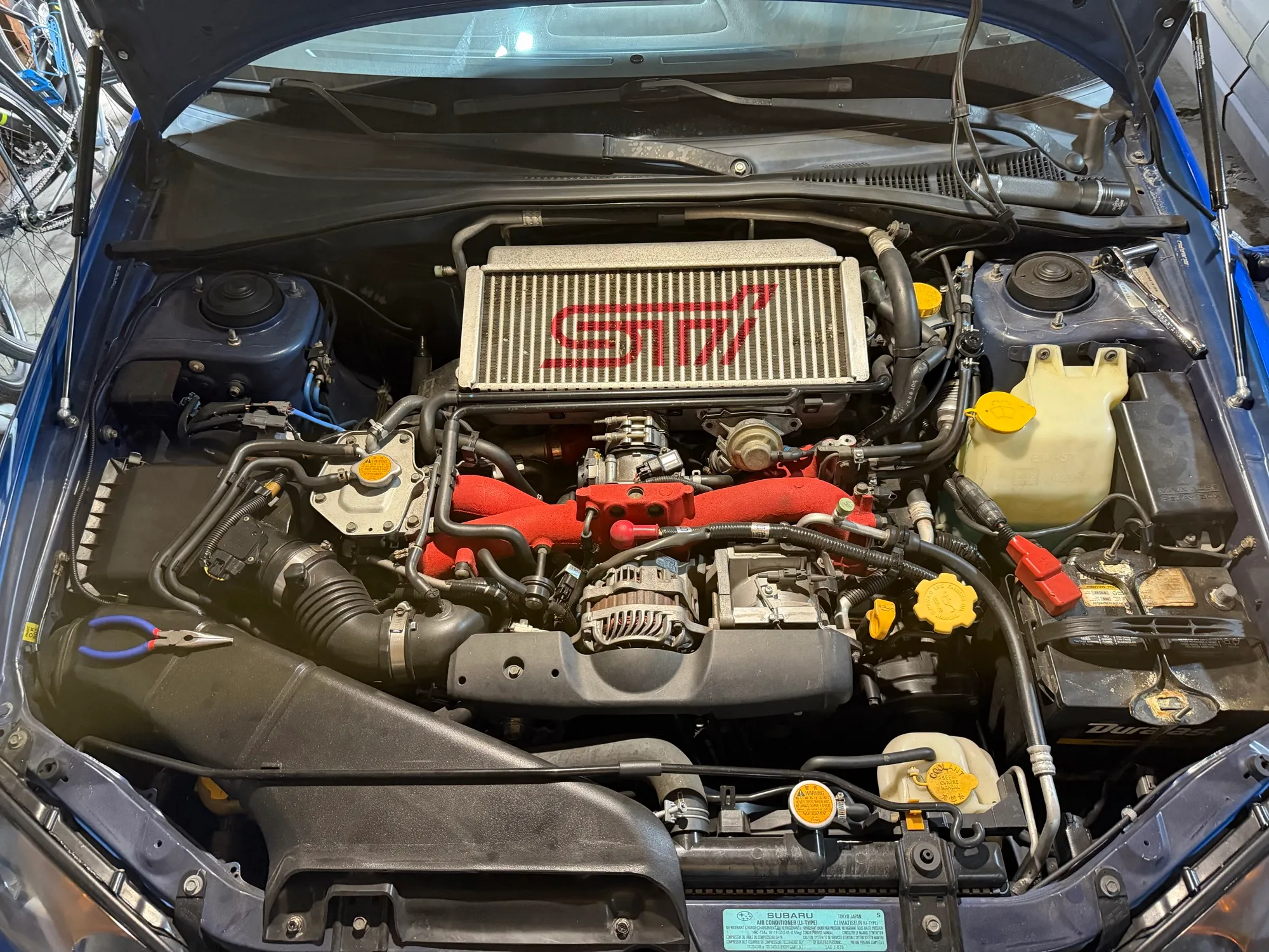 The engine bay of a 2004 Subaru WRX STi with the hood up, the STi top-mount intercooler in place over the red intake manifold.