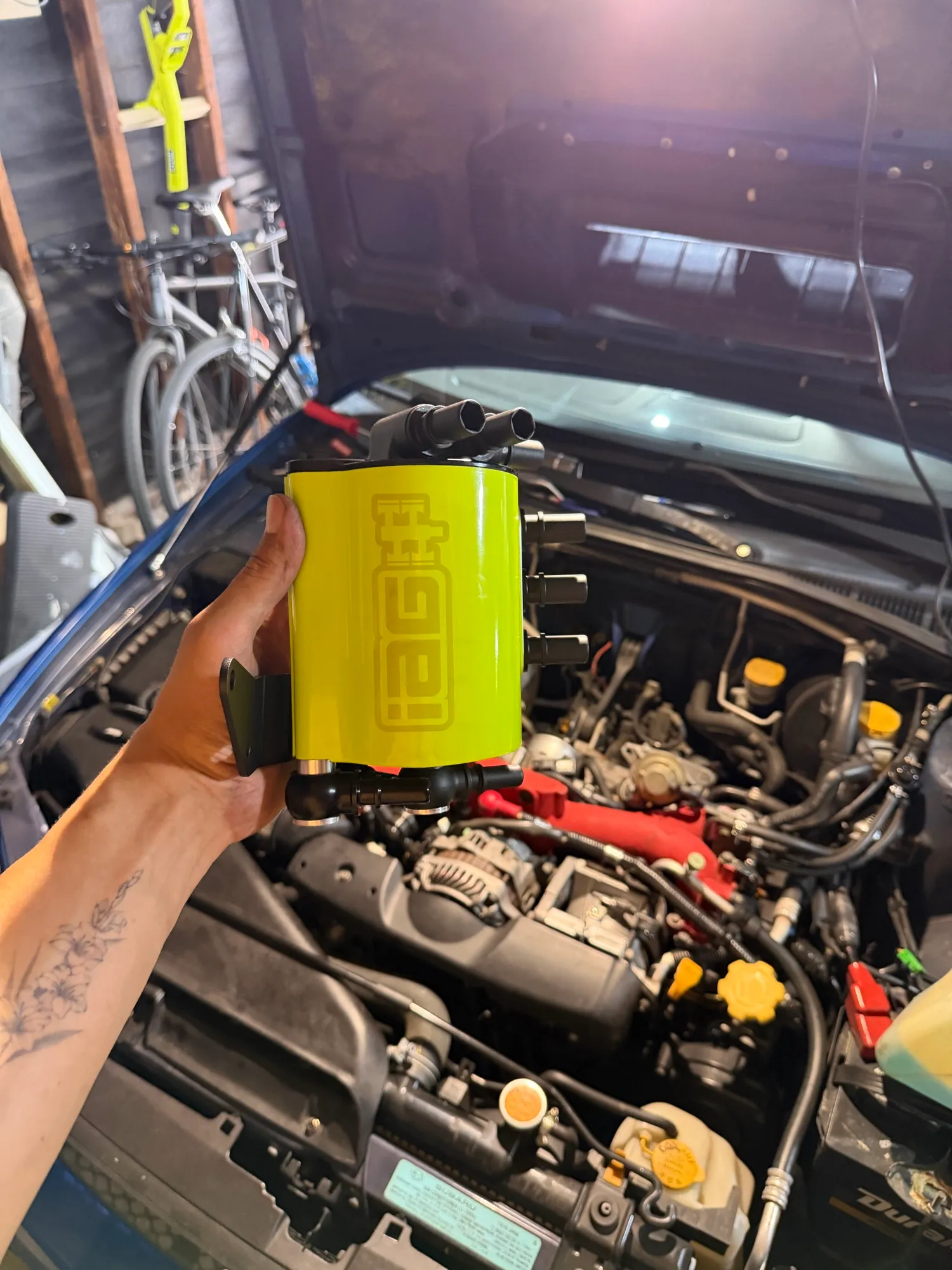 A hand holding the lime green IAG air oil separator can, its fittings pointing up, over the open engine bay.