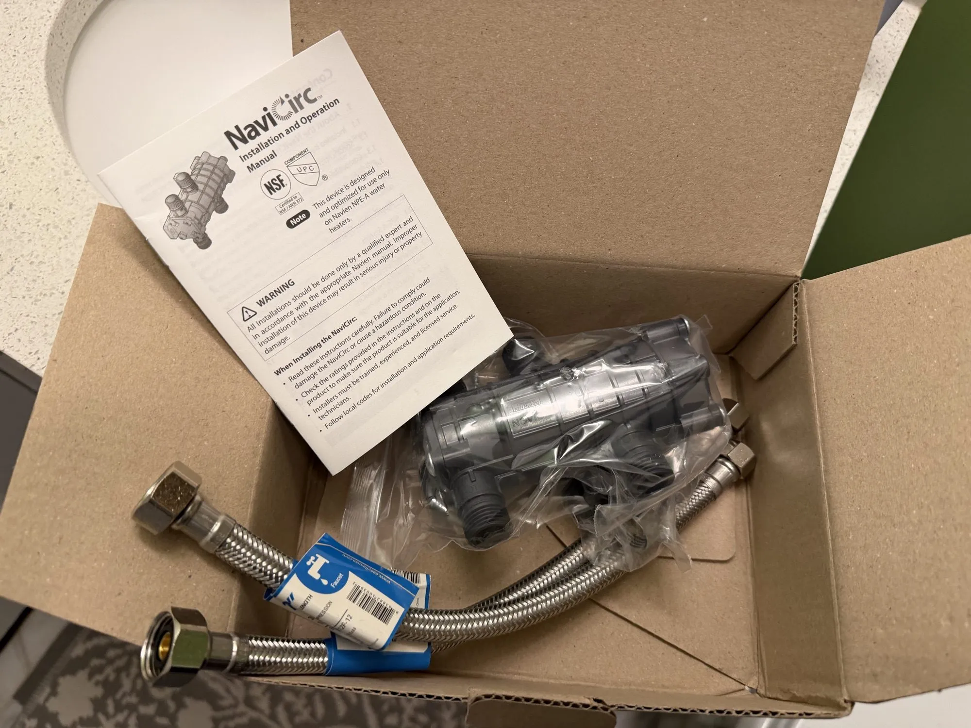 An opened box containing the NaviCirc valve body in plastic wrap, two braided stainless steel supply hoses, and the installation manual.